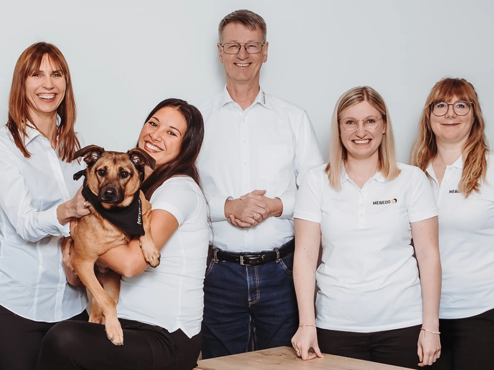 Ein Mann steht in der Mitte. Um ihn herum stehen 4 Frauen, von denen eine einen Hund auf dem Arm hält. Alle lächeln freundlich in die Kamera